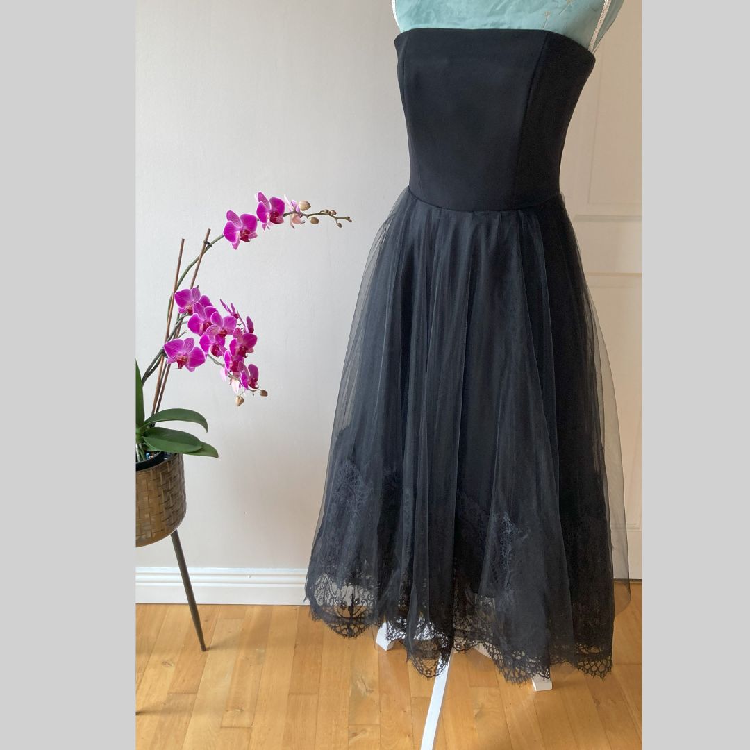 A great fitting strapless dress always creates an iconic and classic look. This statement black gown is composed of a structured boned bodice and a Ballerina style skirt with multiple layers of soft tulle and a French lace edging creating a softness that feels light, luxurious, and romantic.      