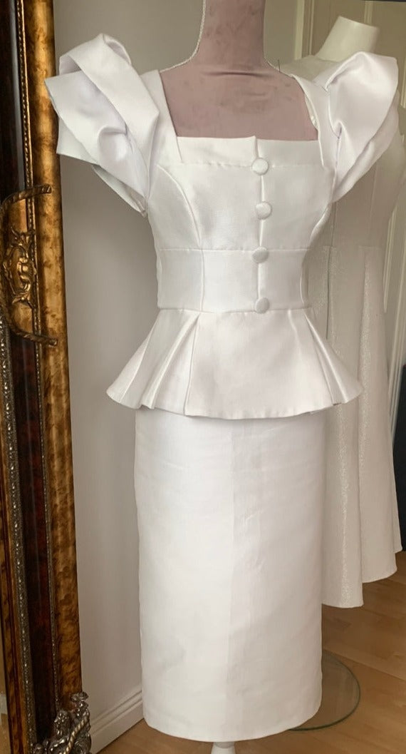 Carmen Sydney Two Piece Skirt & Top suit. Made in a light texture Mikado Ivory Fabric, fully lined in Satin. Featuring button front detail and Dramatic Shoulder in a Layered Rose Effect.